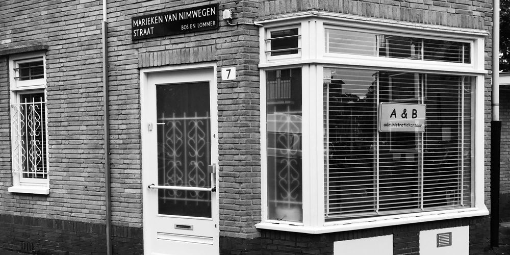 De gevel van het kantoor in Bos en Lommer, in zwart-wit: het straatnaambord, huisnummer 7, de witte voordeur en het raam met het bord van A & B administratiekantoor.